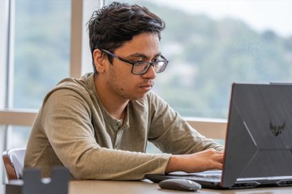 A male student, wearing glasses, is using his laptop.